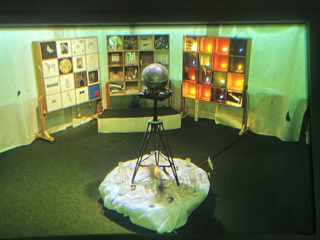Photo of an installation in several parts. Front centre is a silver globe about two feet in diameter, balanced on a spindly stand a few feet tall. Behind are three large wooden frames, each divided into 16 equal squares. The frame on the left has pictures of animals and objects in the squares. The centre one has actual objects and fragments of things. The right hand frame has coloured lightbulbs in the squares. The wall behind is entirely covered with white sheets