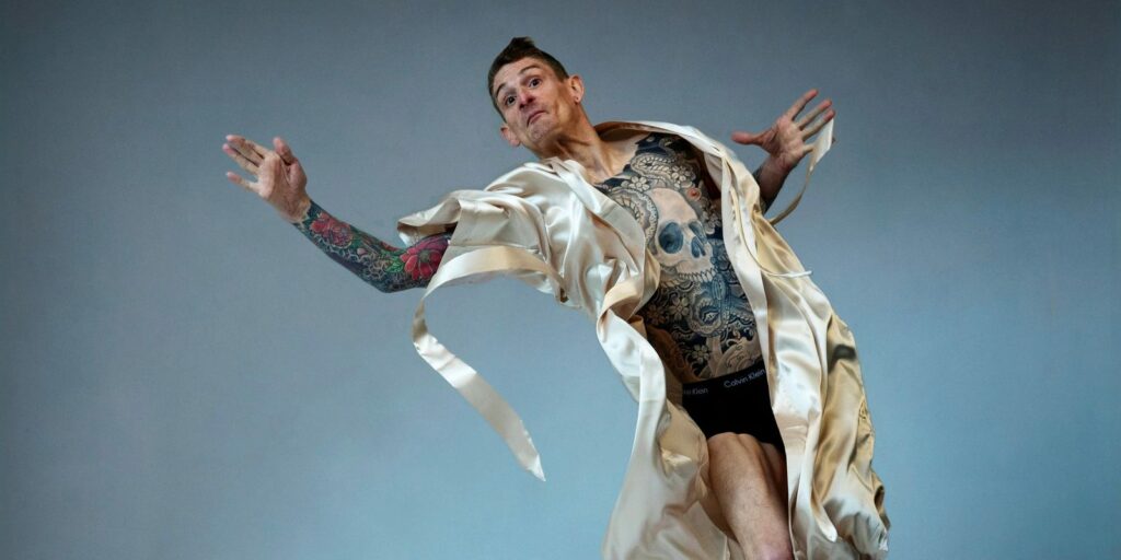 Male presenting figure, the artist Dan Daw, in mid-action pose in mid-air with arms outstretched. There is a mattress covered with a loose sheet beneath them. They are wearing a silk dressing gown that is open, exposing his tattooed skin
