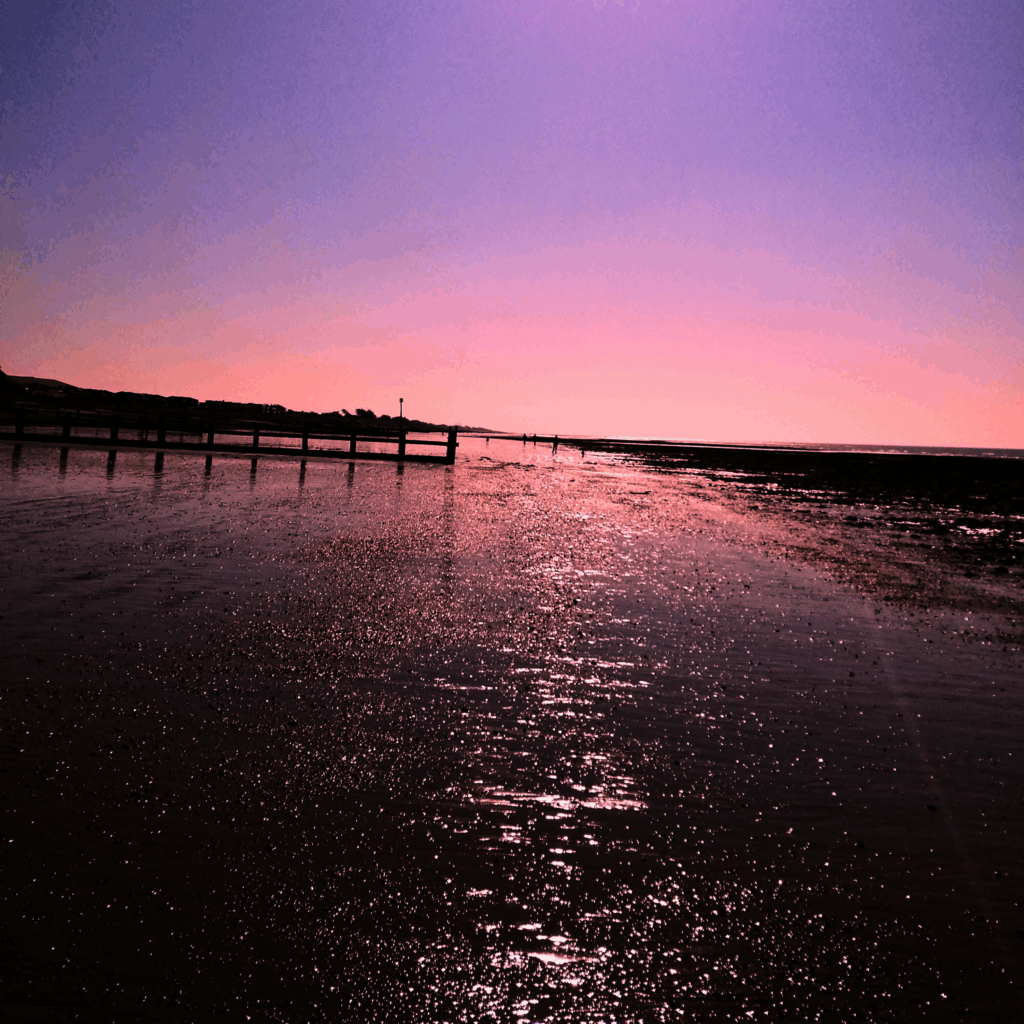 a photo of a beach at low tide. The pink sunset is reflected in the shallow waters, glittering. The silhouette of a wooden sea break is visible on the left in the distance