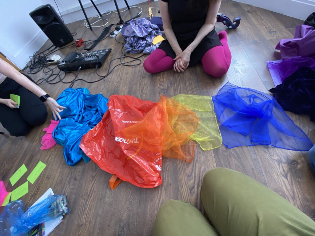 Lowri, Jo and Valeria are sitting on a wooden floor. In front of them, there is a piece of blue fabric, a red plastic bag and sensory scarves (orange, yellow and blue) arranged in a line. There are post-it notes, music equipment and other pieces of fabric around the room.