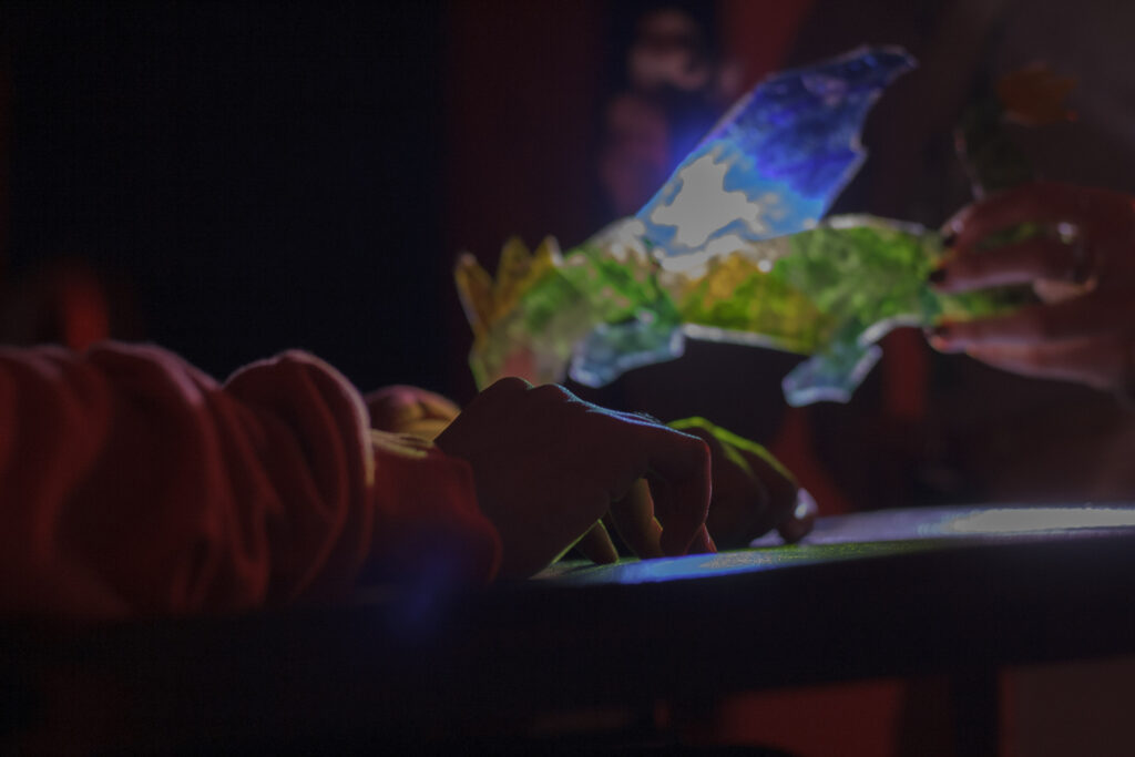 A young person’s hands resting on a tray table. A light shines through a translucent green, blue and yellow dragon puppet, illuminating the young person’s hands and the table in colour.