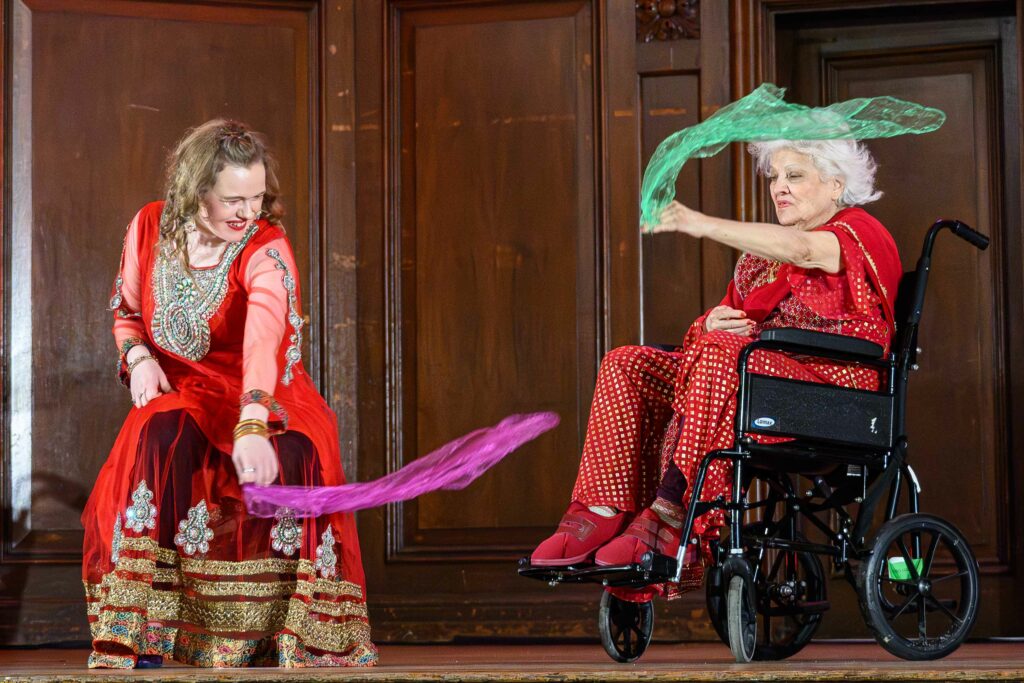 A woman waves a piece of pink tulle. A woman sat in a wheelchair waves a piece of green tulle. They are both wearing red outfits.