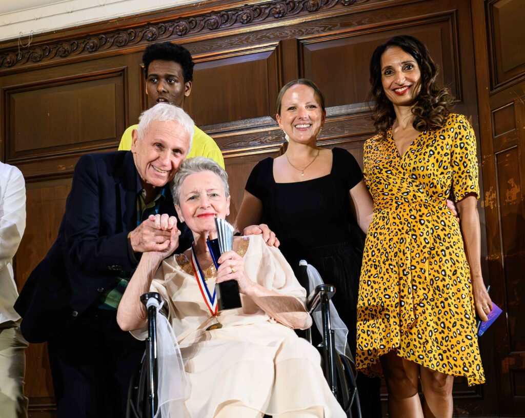 A woman in a cream dress is sat in a wheelchair, holding a trophy and she has a medal around her neck. Ballet dancer Wayne Sleep is stood with his arm around her and holding on of her hands. Two women stand to the side smiling at the camera. 