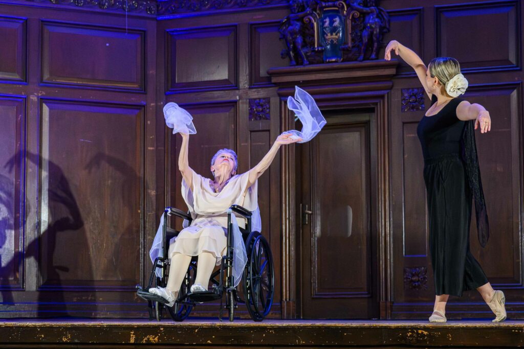 A woman in a cream dress sat in a wheelchair waves white tulle in each hand. A woman to her side wearing a black dress has her arms outstretched.