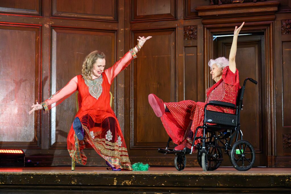 Two women are sat on a chair and a wheelchair. They both have one leg and both arms outstretched. They are both wearing red outfits.