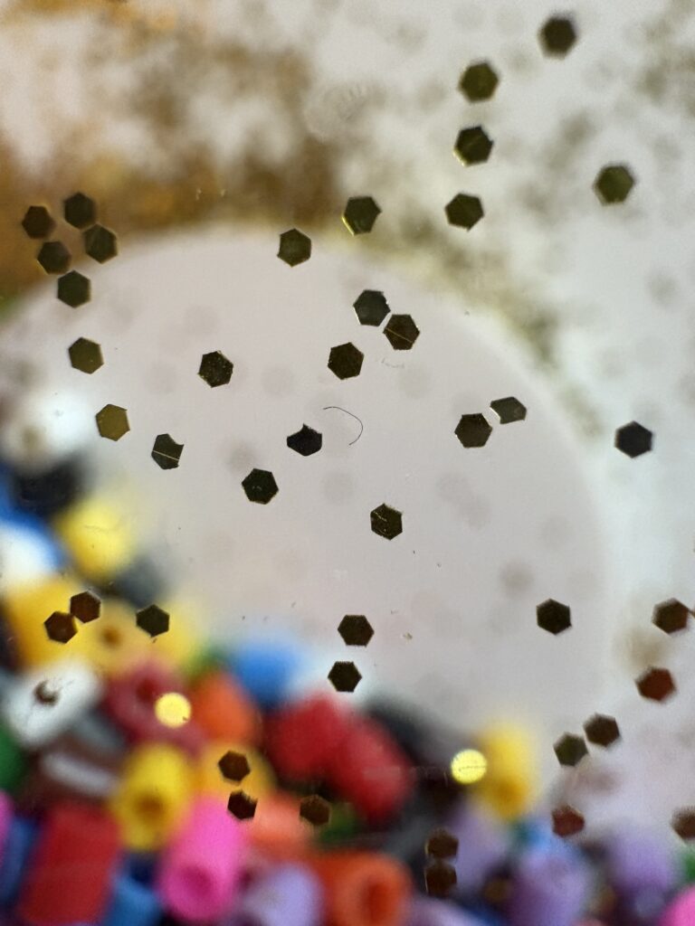 Small multicoloured beads are rendered blurry behind a semi-translucent sheet. Hexagonal sequins are scattered across the surface.