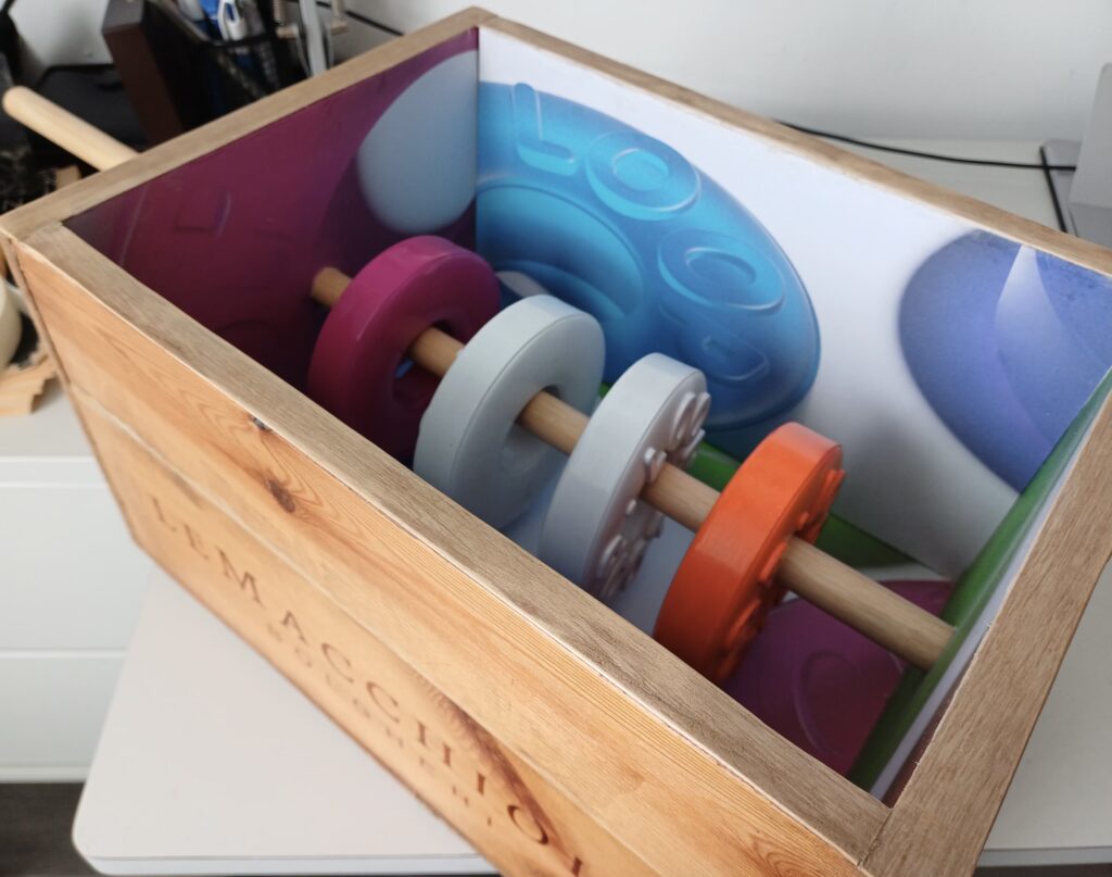 A repurposed wine crate holds sculptural replicas of oversized polo mints, with the letters reading LOOP. A wooden pole through their central holes holds them in place inside the box which is lined with a printed pattern of these loops.
