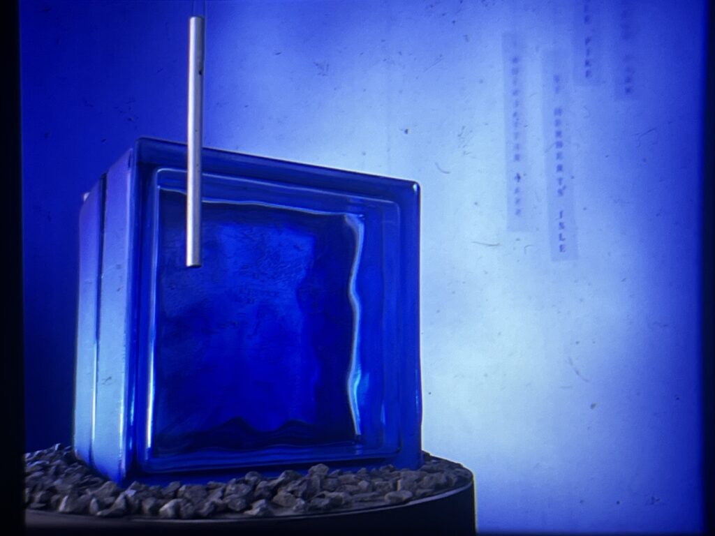 An artwork, a blue box shape with a vertical metal rod in front. In the background are hanging narrow strips with vertical writing. The whole is bathed in blue light