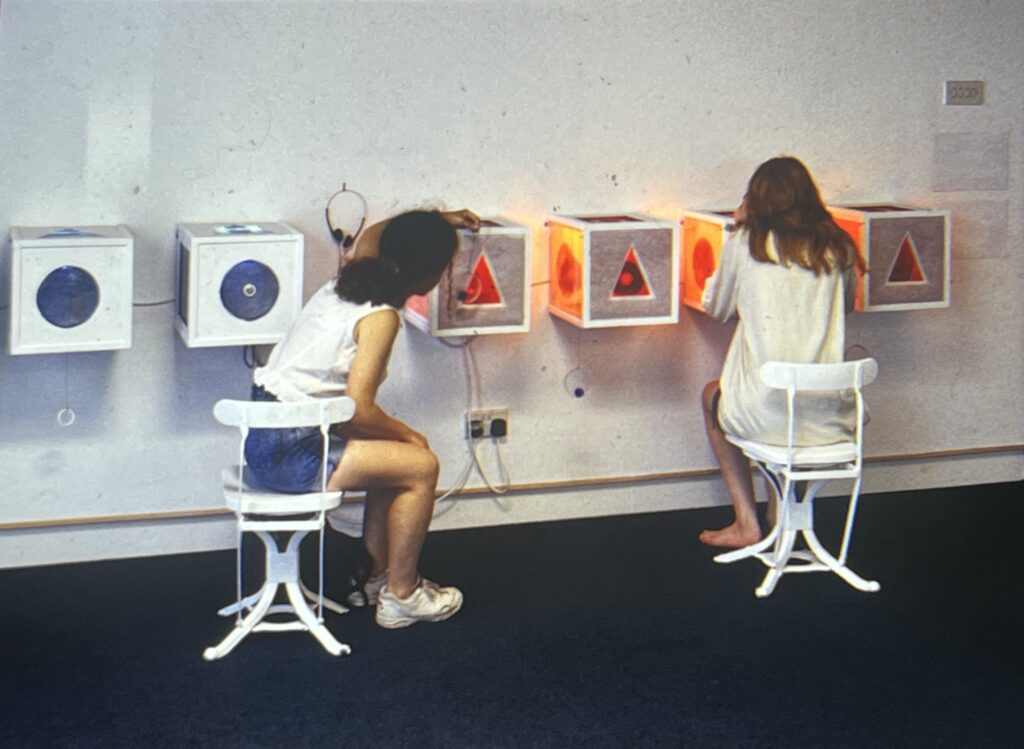 Two seated people reach and peer in to a series of cubes fixed in a horizontal row on a wall. The cubes have triangular and circular holes cut out for the purpose.