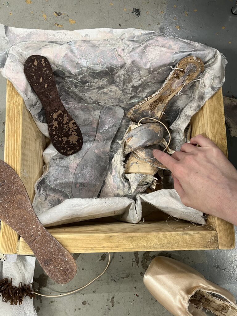 A hand reaches into a wooden box filled with crumpled silky fabrics and the inner sole of a ballet shoe