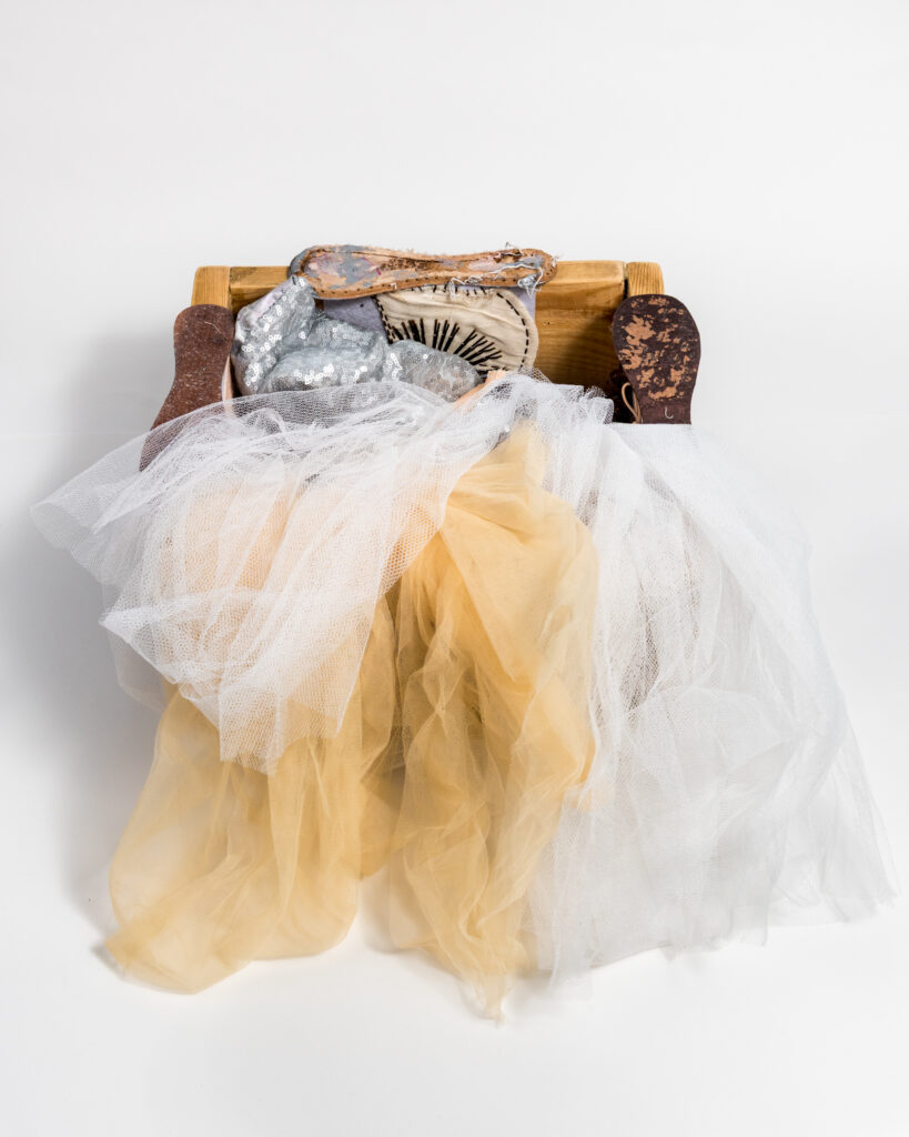 A ballet tutu, deconstructed ballet shoes and other parts of a ballet dancer’s costume spill outof a wooden box