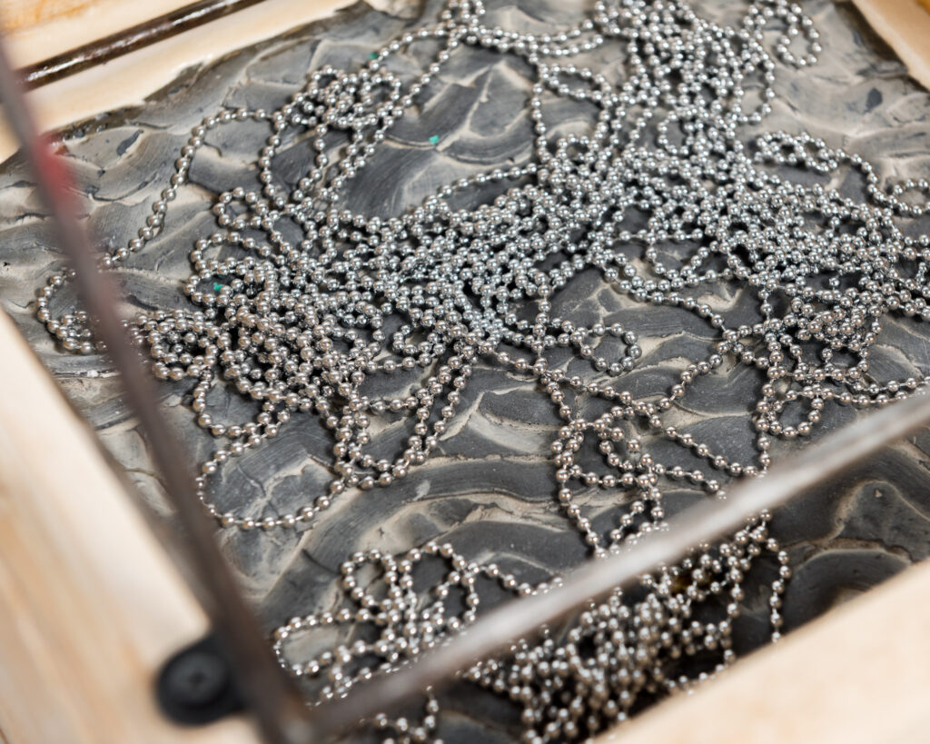 A slinky silver-coloured ball chain rests in wriggly lines on the rippled concrete base of a wooden box. Metal bars have been fixed on top of the box