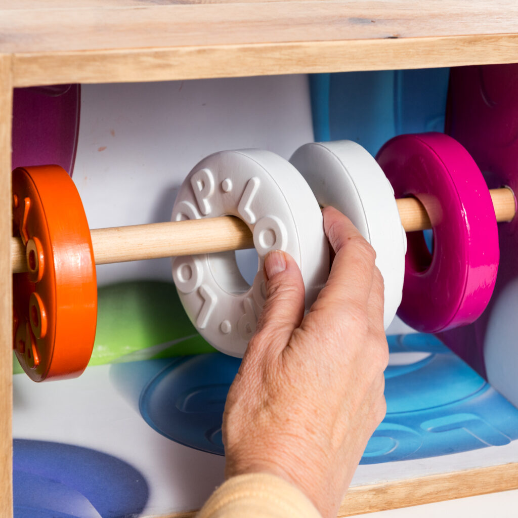 A hand reaches into a wooden box to move one of four rings along a horizontal rod running across. The rings are large replicas of polo mints, although the letters running round read LOOP, and two are white, one pink, one red