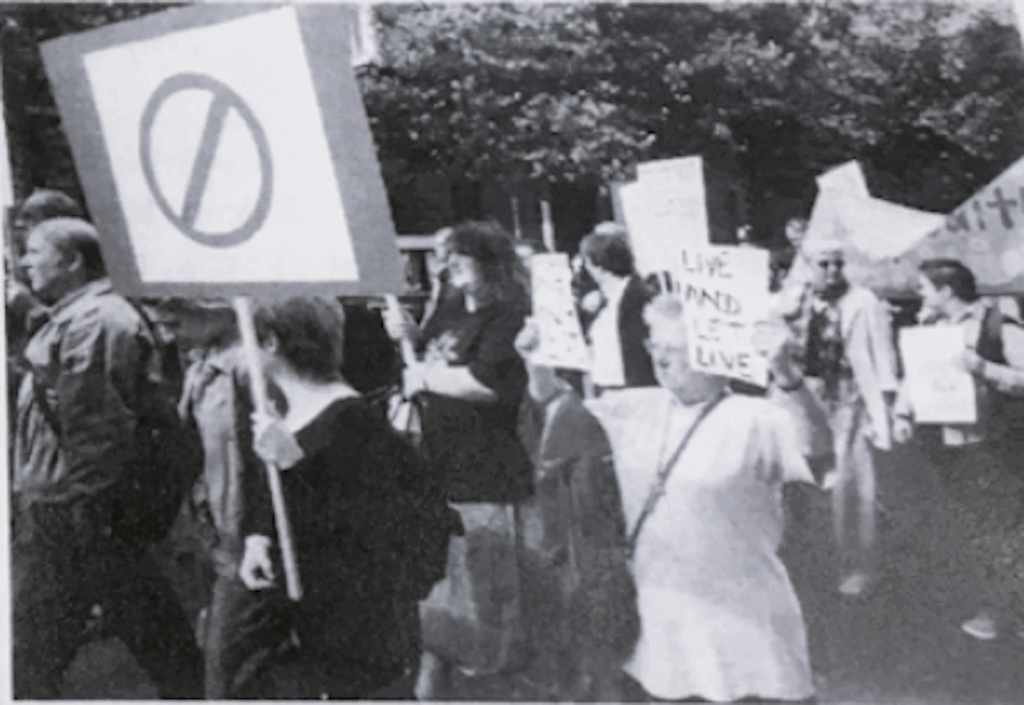 A black and white photo of Audrey in a group at a Mental Health Bill Demo. She is holding up a sign saying 'Live and Let Live.'
