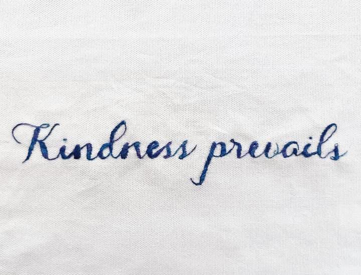 A blue embroidery on white cotton which reads "Kindness prevails"