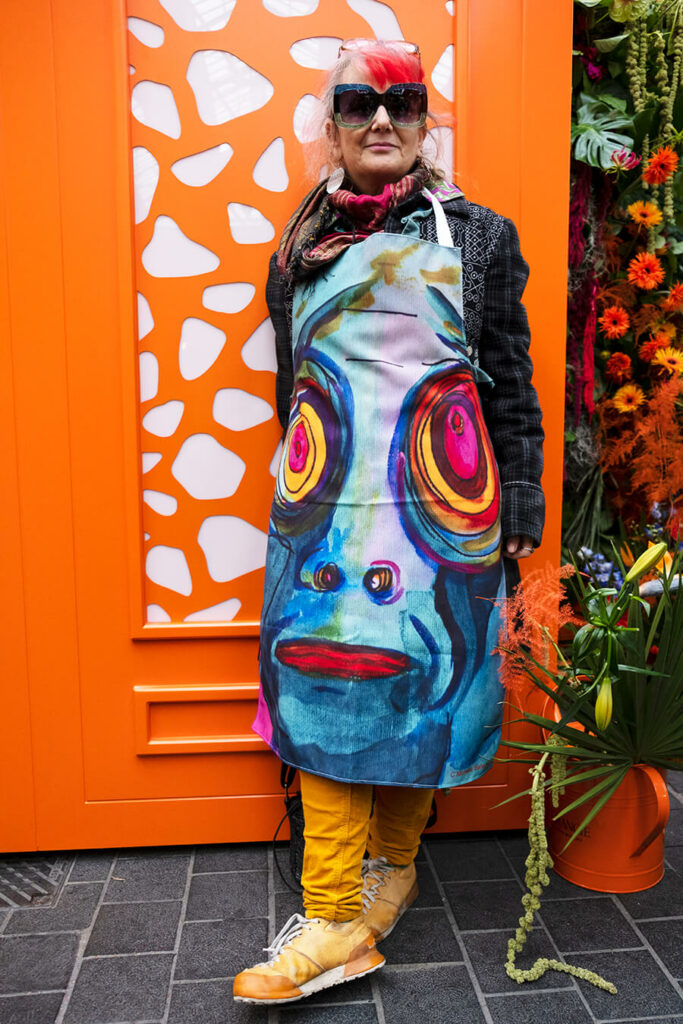 Artist Michelle Baharier stands in front of an orange door wearing sunglasses, a colourful apron with a large face design, yellow pants, and tan shoes, surrounded by orange and red flowers.