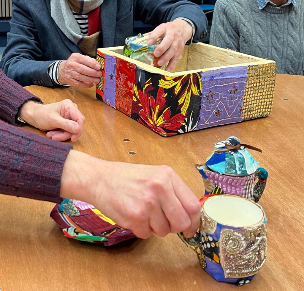 Hands reach across a table to take cups, saucers and milk jug from a wooden box. These are parts of Saba Siddiqui’s A Well Deserved Cuppa and each is covered with a patchwork of patterned fabric