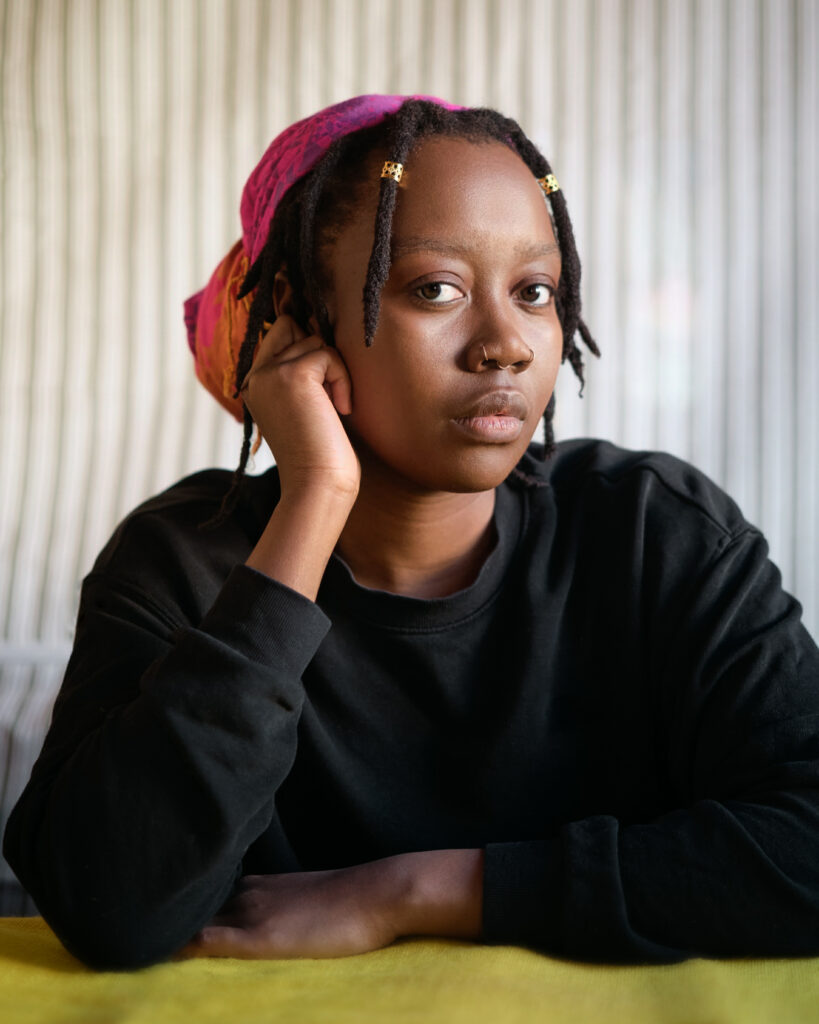 A global majority person with hair in a pink fabric knot and two braids with gold beads framing the front of their face. They are resting the side of their face on one bent arm with with the other in front of them on some green fabric. They are wearing a black sweatshirt and against a pale, striped background