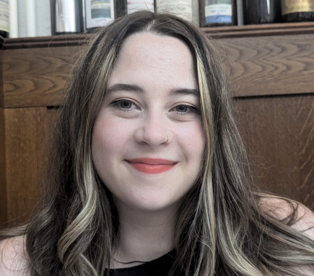 Chloe is a white woman in her early 30s smiles at the camera. She has long brown hair, and is wearing a black top.