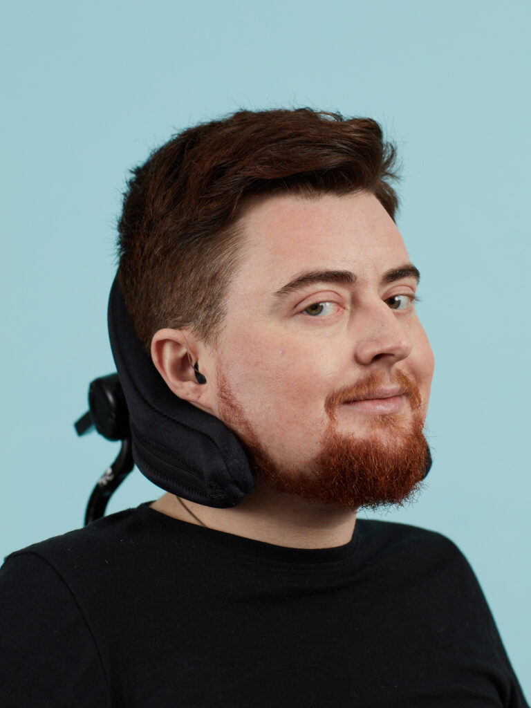 A white electric wheelchair user wearing a black top. They have dark red hair and a red beard. Their head rests on a wheelchair headrest and the background is a studio-turquoise backdrop