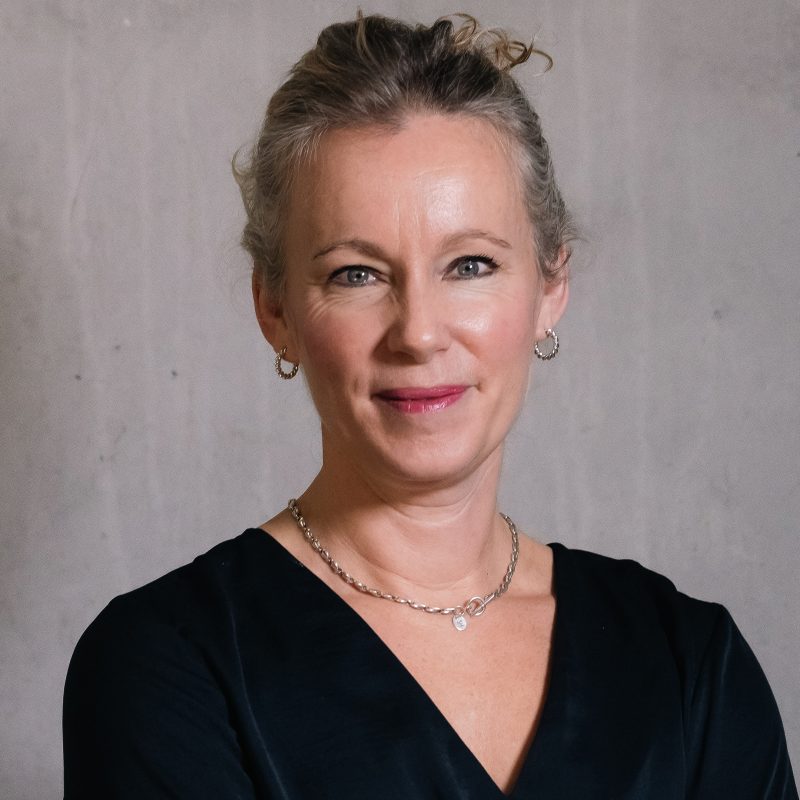 A white woman with brown hair up in a loose pony tail  with silver hooped earring and light pink lipstick wearing a black v-neck top with a chunky silver chain and standing against a grey mottled background