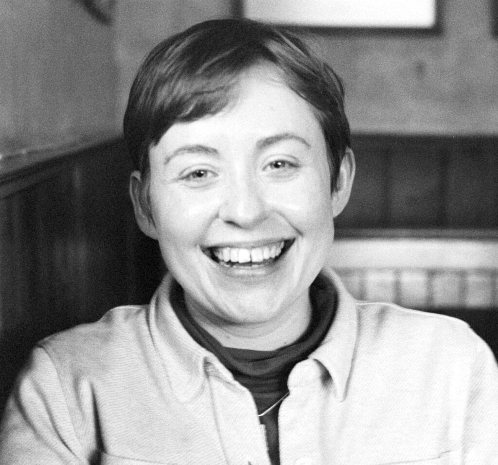 A black and white photo of a white woman in her late twenties with short hair. She is wearing a high-necked top under a thick, buttoned overshirt. She is laughing at the camera and is pictured seated in the interior of her favourite pub in Newcastle-upon-Tyne: the city where she lives and works.
