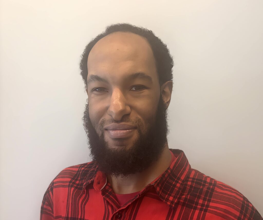 A black man with short black hair and a black beard. He is wearing and black and red check short and standing in front of a white wall