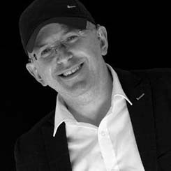 Black-and-white portrait of a smiling man wearing glasses, and a jacket over a light shirt. He is angled slightly toward the camera against a dark background, creating a high-contrast, professional yet relaxed appearance.