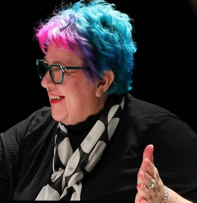 A white woman wearing blue rimmed glasses with blue and pink hair and pink lipstick. She is gesticulating and facing towards the side wearing a scarf with big black and white spots and a black top with lots of silver jewellery. Photo Credit: Black Robin