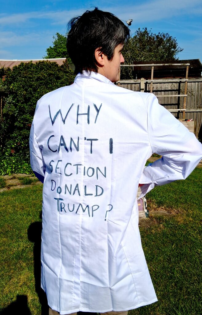Dolly Sen in a doctor's white coat with the text: 'Why Can't I section Donald Trump?' written on the back of it 