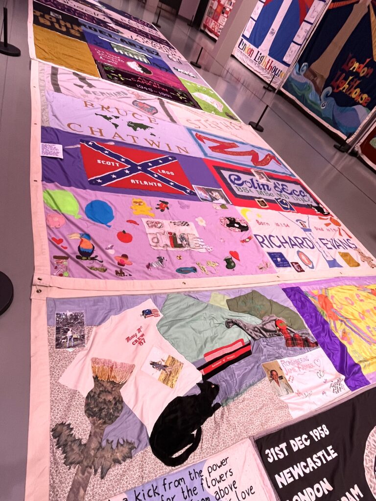 Several hand-made, colourful quilt panels, stitched together in groups, are displayed flat on the floor. Each one is in memory of a person who died from AIDS.