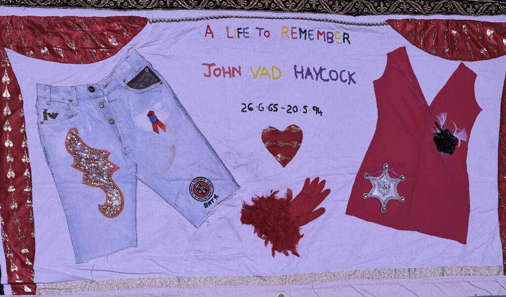 A panel of the AIDS memorial quilt to John Vad Hancock, “A life to remember” embroidered across the top. Actual clothing is attached to this panel - decorated denim shirts on the left, a red t-shirt on the right, plus a feathery red glove between
