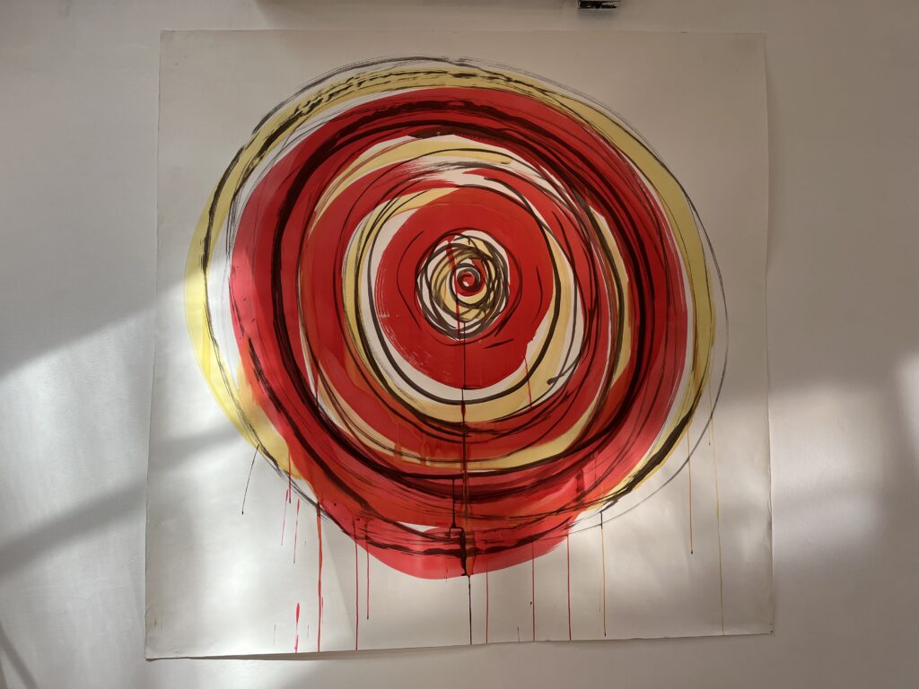 Concentric circles of red and yellow ink in thick brushstrokes on white paper, with thinner black lines adding definition. Shadows from a window fall across the paper