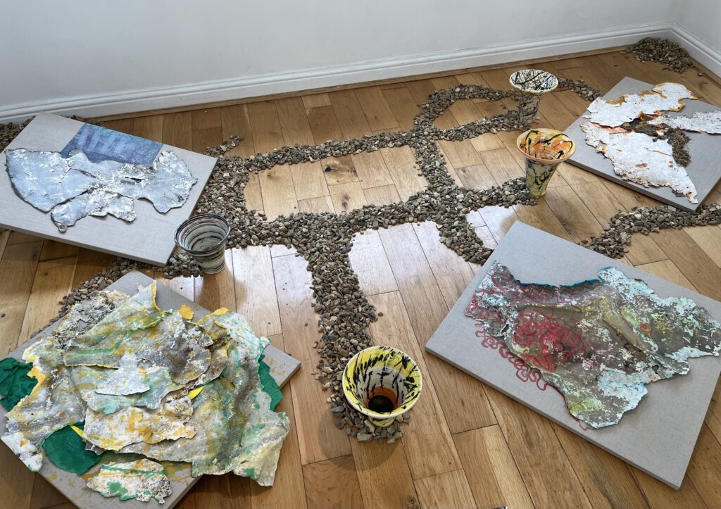 Mixed media artwork arranged on wooden floor: crumpled painted papers, ceramics, and earth