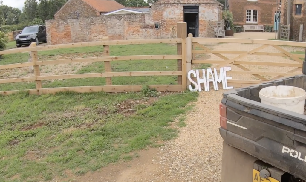 The large cardboard cutout of the word shame propped up against a gated entrance to a house. 