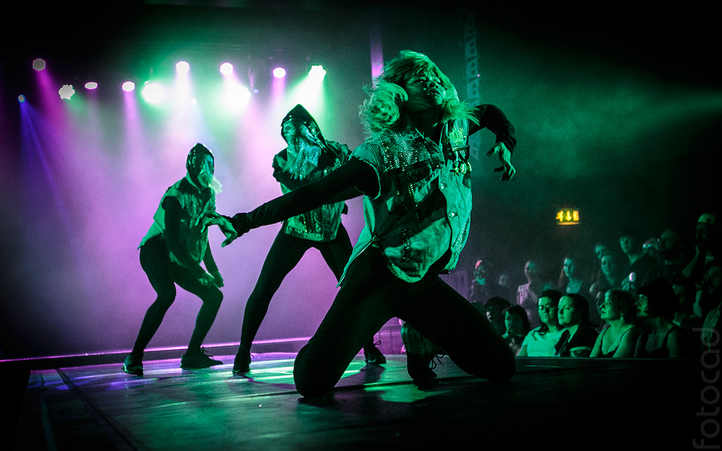 A group of three people dance under purple and green lighting on a dark stage while an audience watch on. They are wearing black leggings and cut off denim jackets with diamonte. The two dancers at the back are wearing lace face coverings and Ariel Fung the dancer at the front has long blonde hair and kneels on the stage doing a wide arm gesture.