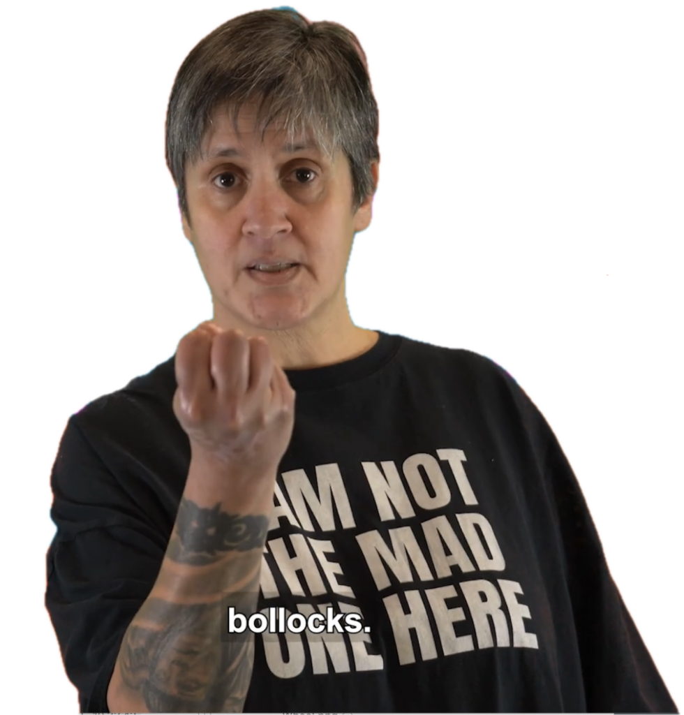 A film still - Dolly stands with a clenched fist towards the camera. She is wearing a black T-shirt with white text on that reads: I am not the mad one here. Captions reads: Bollocks.